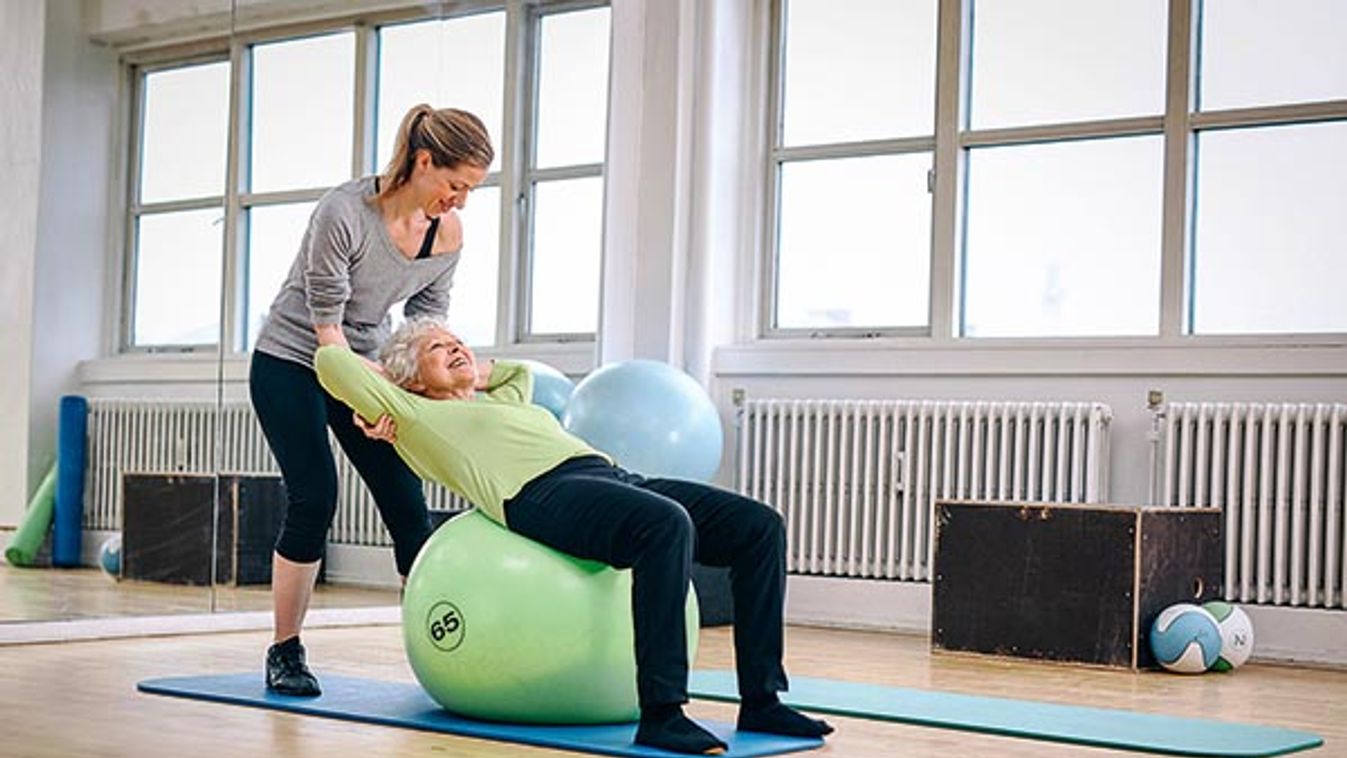 Female coach helping senior woman exercising in gym. Elder woman working out on pilates ball at health club being assisted by he